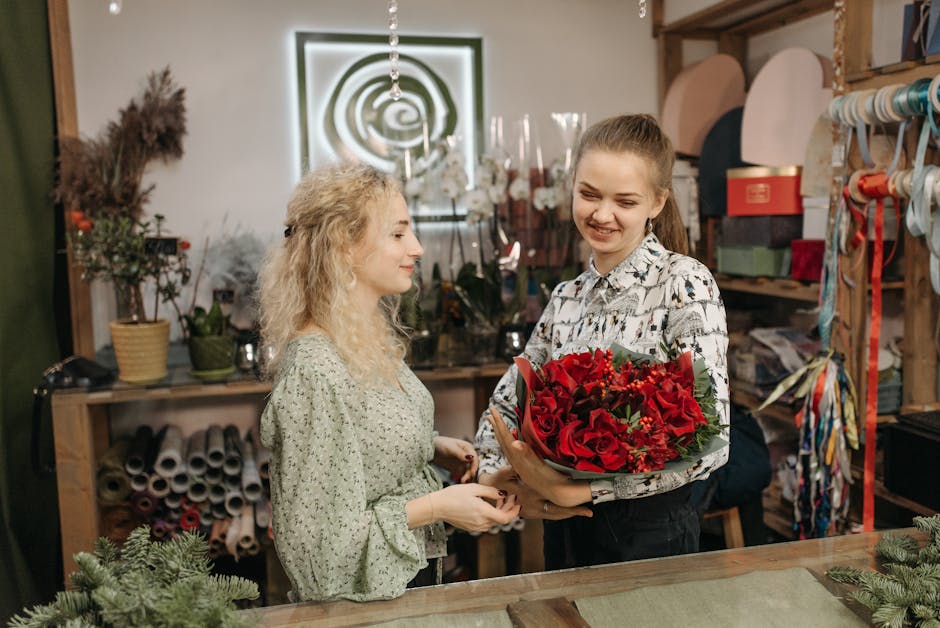 A florist with long, light brown hair tied back, smiling as she presents a large bouquet of vibrant red roses, lilies, and other mixed flowers wrapped in brown kraft paper, to a customer with curly bl
