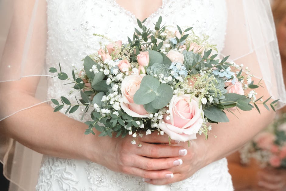 A bride holding a delicate bouquet of fresh flowers composed of soft pink roses, pale blue hydrangeas, sprigs of baby's breath, and eucalyptus leaves in shades of green. The bouquet is arranged in a c