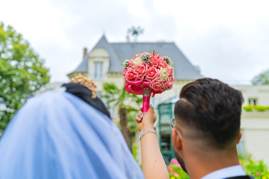 A close-up view of a floral bouquet held by a person, featuring a tight arrangement of pink and cream roses with small white filler flowers and lush green foliage, wrapped with a bright pink ribbon ha