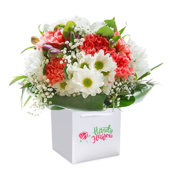A celebration of local Perivale charm, this radiant floral arrangement is presented within a chic white gift bag accented by sturdy ribbon handles, set against a flawless white photographic backdrop. The bouquet's highlight is the wealth of coral and fleshy-pink carnations, their ruffled, layered petals offering soft volume and gentle elegance. Vibrant red carnations peek through the palette, adding striking depth and an emotional undertone perfect for moments of genuine sentiment. Scattered with care, bright white daisies with distinctive chartreuse centers introduce clarity and light, brightening the bouquet with an uplifting, everyday joy reminiscent of sunny afternoons by the Grand Union Canal. Baby's breath, in delicate clusters, gently weaves through, lending a cloudlike softness that softens the arrangement's edges. Unopened flower buds tinged with purple nestle toward the side, presenting a subtle nod to the cycles of growth and renewal seen each season in Perivale's vibrant community gardens. Encasing the flowers, broad, dark green foliage envelops the base, forming an elegant collar which keeps the arrangement full and structured. The bouquet's rounded silhouette and careful composition underline a sense of generosity and celebration, perfect for gifting during birthdays, anniversaries, and heartfelt occasions celebrated across Perivale. Floristry craftsmanship is evident in every detail, creating not just an arrangement, but a memorable floral experience made for this unique West London neighbourhood.