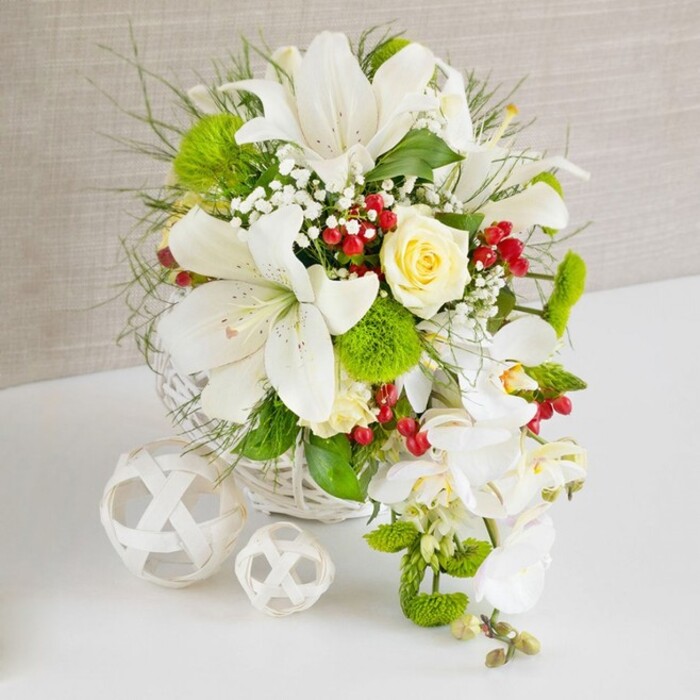 A meticulously arranged white and green floral centerpiece is displayed on a clean white surface, evoking the polished elegance of a Perivale celebration set near the green spaces around Horsenden Hill. The bouquet is cradled in a round white wicker basket, whose smooth, interwoven strands offer a subtle texture and a handcrafted feel. At the heart of the arrangement, several large white Oriental lilies rise proudly, their trumpet-shaped petals open and luminous, revealing fine speckling and yellowish stamens dusted with pollen. Just above and among them, a single cream or pale-yellow rose is positioned as a soft focal point, its spiral of petals tightly layered, suggesting freshness and expert handling. From the right-hand side of the basket, a cascade of pure white phalaenopsis orchids descends, each bloom elegantly spaced along its arching stem, creating a waterfall or teardrop effect that adds graceful movement and a distinctly bridal touch. Surrounding these focal flowers, a rich array of greenery fills the basket: slender, wispy grasses and feathery fronds add lightness, while broader, glossy leaves give substance and structure. Bright green pom-pom chrysanthemums or mossy dianthus balls dot the arrangement, introducing playful, tactile spheres of colour that contrast beautifully with the smooth lily and orchid petals. Fine sprays of white baby's breath lace through the design like delicate mist, softening the transitions between blooms. Scattered among the whites and greens, clusters of ruby-red berries offer bold, jewel-like accents that bring warmth and seasonal richness. To the left on the table, two white decorative spheres of different sizes, crafted from interlaced material, rest quietly, their open latticework echoing the wicker basket and completing the composition. The scene feels calm, sophisticated, and celebratory, ideal for a wedding table, engagement party, or refined family gathering in Perivale, where fresh, thoughtfully designed flowers set the tone.