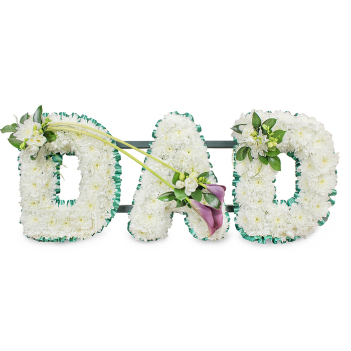 This photograph captures a refined, personalised funeral floral display spelling out "DAD" in bold capital letters, ideal for families in Perivale wishing to honour a much-loved father. The three letters are constructed on a sturdy, horizontal frame and designed to stand upright, which is subtly indicated by dark supporting bars visible behind the flowers. Every surface of each letter is densely packed with pure white chrysanthemums or similar button blooms, placed so closely that the base cannot be seen. The result is a deeply cushioned, cloud-like texture that feels serene and comforting, perfect for a condolence tribute at a church or crematorium serving the Perivale area. Around the outer edges of the letters, a ruffled emerald green ribbon has been meticulously pinned, creating a clean outline and bringing a fresh, natural contrast, similar to the vibrant greens of nearby golf courses and open spaces. The first "D" carries a small floral accent on its upper left curve: a gentle cluster of white freesia and glossy foliage, arranged to sit gracefully atop the chrysanthemum bed. Two long, arching green stems extend from this area, curving diagonally across towards the central "A" and forming a visual bridge between the letters. Nestled near the lower side of the "A" is the main focal spray, where two striking deep purple or maroon calla lilies are nestled among smaller white blossoms and leaves. The calla lilies, with their smooth, funnel-like petals, offer a rich, dignified note of colour against the sea of white. On the final "D," the upper right edge is adorned with a smaller but matching cluster of white freesia and foliage, tying the entire design together. The clean, white background isolates the tribute, allowing its message of love, remembrance, and respect to stand out clearly for any farewell gathering in Perivale.