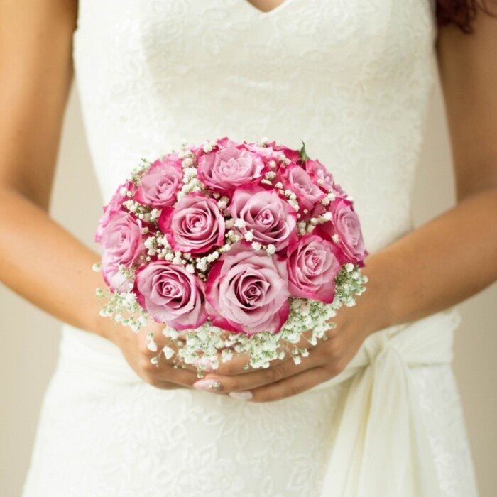 The image captures a Perivale bride's loving hands as they cradle an exquisitely hand-tied round bouquet-a tribute to the area's enduring romance and floral craftsmanship. The bouquet, striking in its compact shape, features around twelve lush roses, each displaying a subtle yet striking gradient: the innermost petals shimmer with soft, pastel pink, while the edges deepen into radiant fuchsia and bold magenta, recalling the vibrant blooms in Perivale's neighbourhood gardens. Interwoven among the roses, countless tiny blossoms of baby's breath (gypsophila) provide billowy, cloud-like contrast, lending texture and a gentle, classic feel that resonates with Perivale's timeless venues. The bride's hands, tanned and lovingly manicured with a soft, light polish, wrap around the bouquet above the intricate floral lacework of her sleeveless wedding gown. A delicate flowing sash falls gracefully to the right, further enhancing the scene's layered textures. Bathed in soft natural light with a muted neutral backdrop, every flower, petal, and hint of greenery stands out, inviting onlookers to share in the joyful hope of a Perivale wedding day. This image is a perfect expression of the harmony between nature and tradition, local artistry, and the heartfelt anticipation of two lives joined in love.