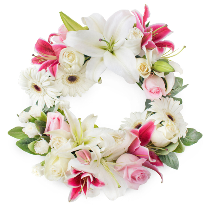 This open circular sympathy wreath is shown against a white background, an elegant ring of blooms that combines white, soft pink, and vivid magenta to create a calm yet emotionally resonant tribute. The eye is initially drawn to the large white lilies spaced around the circle, each bloom fully open, with thick, satin-like petals that arch outward from fresh green centres dusted with golden pollen. They convey a sense of purity and peace, echoing the quiet, reflective atmosphere of Perivale's leafy residential streets on a still afternoon. Nestled between them are deep pink and fuchsia lilies, their starry petals and gentle speckles offering pockets of intense colour that speak of cherished memories and enduring love. Clusters of crisp white gerbera daisies, each with a distinct dark centre, add rhythm and brightness, their layered petals radiating like small, contained bursts of light. Interspersed are soft pink roses, some just budding, others loosely opened, suggesting tenderness, gratitude, and affection, while creamy white roses in fuller bloom bring volume and a classic, almost timeless character to the piece. Smaller white filler flowers, such as petite carnations or spray chrysanthemums, are woven into every space, softening transitions between the larger blooms and ensuring the wreath feels complete and abundant. A lush collar of green foliage frames the flowers, with overlapping leaves and subtle variations in tone giving depth and structure, reinforcing the symbolism of the unbroken circle and the continuity of life and love. The arrangement gives the impression of having been crafted by a skilled Perivale florist, ideal for funerals, memorial services, or celebration-of-life occasions where families want a floral tribute that is both visually graceful and quietly consoling.