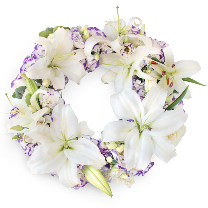 This beautifully crafted circular funeral wreath is shown against a bright white background, allowing every nuance of its white and purple palette to register clearly, as if lit by soft daylight filtering through a chapel window in Perivale. The ring is generously composed of large white Oriental lilies, whose broad, velvety petals open in tranquil arcs. Many of these lilies are at their fullest, revealing a slightly green throat and gracefully extended stamens ending in golden-brown anthers, while a few buds remain partially closed, waiting to unfold. Their serene faces lend an air of purity and spiritual calm, echoing the traditional use of lilies to honour the soul's passage. Interlaced with these lilies are ruffled lisianthus-style flowers, their white petals edged softly in lilac and purple, reminiscent of the gentle evening tones you might glimpse over the Greenford Road from Perivale. These purple fringes add both visual depth and emotional resonance, suggesting reflection, dignity, and quiet remembrance. Deeper within the wreath, small lavender-toned fillers and occasional cream buds subtly break up the surfaces, providing layers that would feel plush and comforting under the fingertips. A base of dark green foliage and fine, eucalyptus-like sprigs forms the hidden backbone of the design, their cool greens adding structure and contrast without overpowering the blooms. The overall circle is full and unbroken, meticulously balanced so that no area appears denser than another. Crafted with a sensitivity to the needs of grieving families in Perivale, this wreath serves as a dignified tribute for funerals and memorial gatherings, a floral expression of love, peace, and the continuity of memory that surrounds a life well lived.