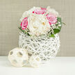 In this serene floral composition, a lush, dome-shaped bouquet of ivory peonies and deep fuchsia-pink roses is showcased in a modern, openwork white vase, reminiscent of the contemporary homes and wedding venues around Perivale. The peonies form the heart of the arrangement, their generously layered petals unfolding in soft, creamy whorls that look almost weightless, conjuring thoughts of a classic garden perfume. Encircling them, the roses provide a vivid contrast, with petals that shift from warm blush near the centre to intense pink at the edges, bringing a sense of passion and joy. Threaded between these larger blooms, clusters of baby's breath introduce a fine, lacy texture, each tiny white flower catching the light like drifting snowflakes and creating a gentle halo effect around the bouquet. At the base, rich green leaves fan out gracefully, polished and slightly curved, giving the piece a balanced, structured finish that feels both natural and meticulously crafted.
The bouquet is nestled into a distinctive, spherical vase made of thin white strands that interweave and cross, forming a loose, lattice-like shell that calls to mind woven rattan or twigs painted in crisp white. This airy structure allows glimpses of stems and interior shadows, adding depth and a sense of movement, as though the flowers are cradled in a sculptural nest. Resting on the smooth, bright white tabletop are two decorative spheres woven from pale, straw-coloured material, one larger and one smaller, echoing the vase's texture and extending its organic, artisanal theme.
Behind the arrangement lies a neutral beige background with a subtle linen-like texture, keeping the eye focused on the bouquet and its interplay of colour and form. The lighting is bright yet softly diffused, ensuring that each petal, leaf edge, and woven strand is gently illuminated without harsh glare. The result is a display that feels refined and tranquil, ideal for a Perivale reception, a special anniversary meal at home, or any moment that calls for quiet, romantic beauty.