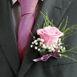 Pinned to a dark charcoal suit, this boutonniere features a single blush-pink rose that appears almost luminous, as though caught in gentle afternoon light filtering through trees in Perivale. The rose is the clear focal point: its petals unfurl in a precise, graceful spiral, each layer soft and velvety, with a delicate transition from pale pink at the edges to a slightly warmer tone near the centre. Around the bloom, airy white gypsophila forms a light, cloudlike frame, each tiny clustered flower adding intricate texture that softens the contrast with the suit. Slender green foliage-fine fern fronds or similar leaves-peeks out from beneath, extending subtly outward, giving the boutonniere a sense of movement and natural ease, reminiscent of a gentle breeze over Perivale's open fields. The stems are carefully bound with a narrow satin ribbon in a coordinating shade of mauve or dusty pink, wrapped smoothly and tied with a discreet bow that showcases the florist's attention to detail. Light highlights the delicate curves of the rose petals and lends a faint sheen to the ribbon, while faint shadows emphasise the boutonniere's sculpted shape. This arrangement would be perfectly suited to a wedding or formal celebration in Perivale, Greenford, or nearby Ealing, whether at a local church or a modern venue. It conveys tenderness, thoughtfulness, and a sense of polished simplicity-an elegant floral accent meant to accompany important vows, cherished photographs, and joyful gatherings that friends and family will remember.