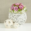 A serene, studio-style scene shows a contemporary floral centerpiece that would suit a Perivale wedding reception or an intimate dinner near the open spaces of Perivale Park. At the center is a white, spherical vase made from slim rattan or twig-like strands intricately woven into an open, airy lattice. The container's round form and visible gaps give it a light, sculptural presence on the smooth white tabletop. Rising from this base is a perfectly rounded dome of fresh flowers, dominated by roses in tender shades of pink and soft lavender, their petals unfurling in lush spirals. Some blooms display deeper pink along their edges, introducing a gentle gradient of color. Tucked between the roses, bright green button chrysanthemums punctuate the bouquet with structured, spherical shapes and a burst of vivid green that enlivens the pastel palette. At the lower edge, delicate white filler flowers wind in and out among the larger blossoms, creating a soft, almost frothy border that slightly overlaps the vase. To the left side of the arrangement, two decorative woven balls in pale, natural wood tones rest on the same white surface-one larger, one smaller-each crafted with an open weave that echoes the texture of the vase and adds visual balance. The background is a softly blurred field of light beige or tan fabric, reminiscent of linen drapery, which contributes warmth and subtle texture without drawing attention away from the flowers. Soft, even lighting bathes the scene, highlighting the roses' velvety surfaces, the crisp green mums, and the intricate wicker work, conveying an atmosphere of calm refinement ideal for illustrating wedding table arrangements and modern floral design in Perivale.